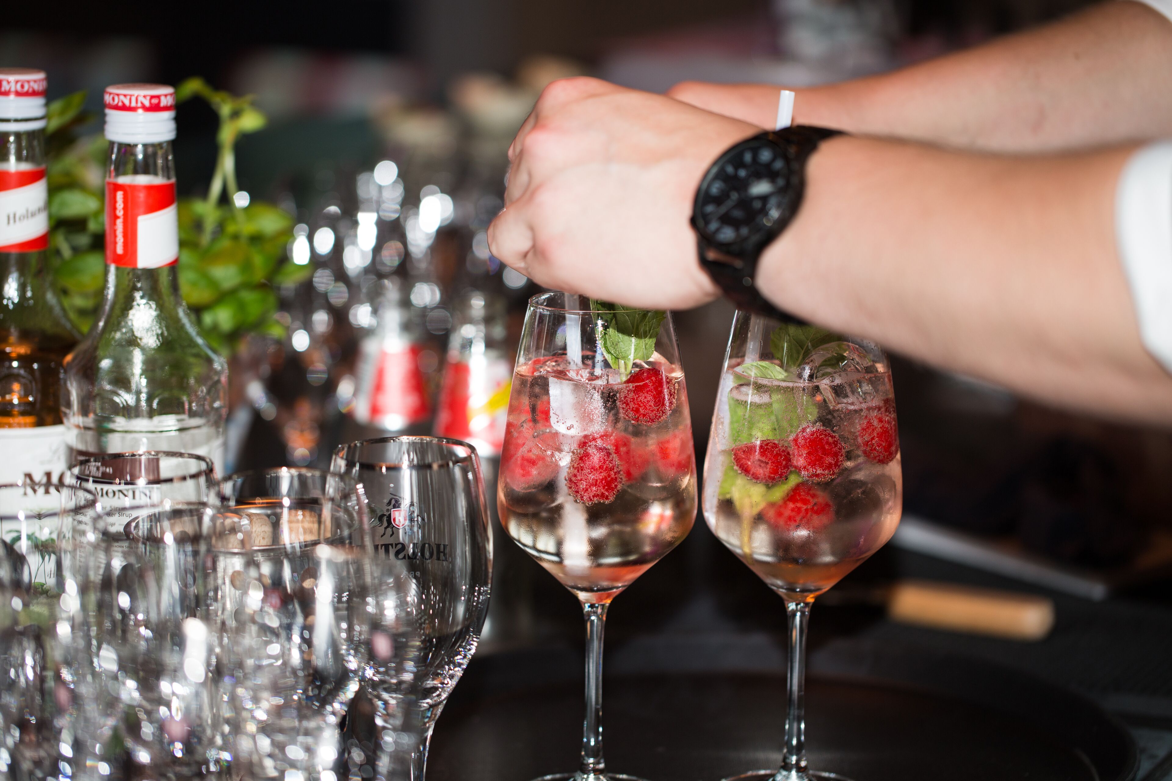 A bar setting with multiple glasses of a strawberry drink, a bottle of liquor, and a watch on a wrist.