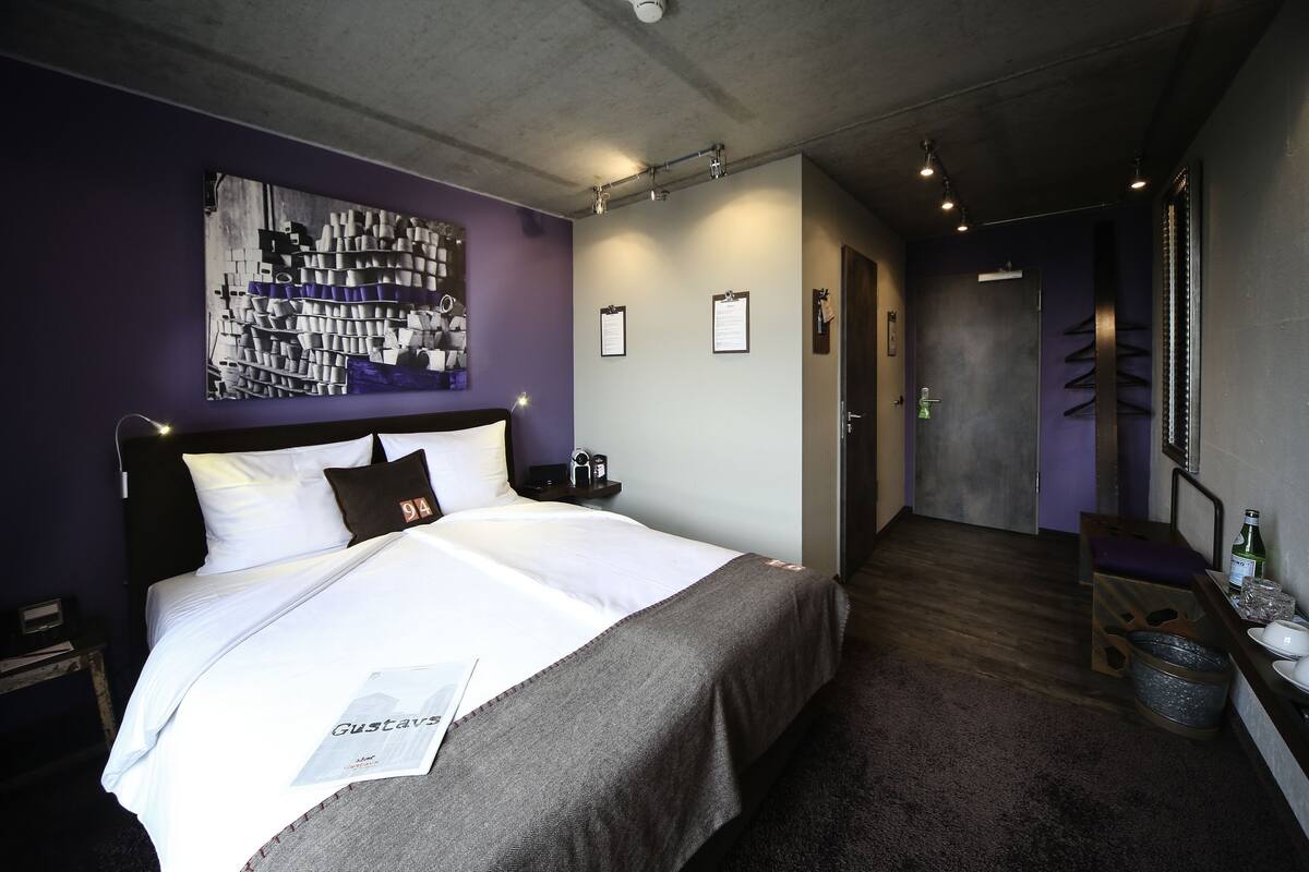 Quarto de hotel moderno com uma cama grande, cabeceira, obra de arte na parede, mesa de cabeceira com abajur e uma pequena escrivaninha com cadeira.
