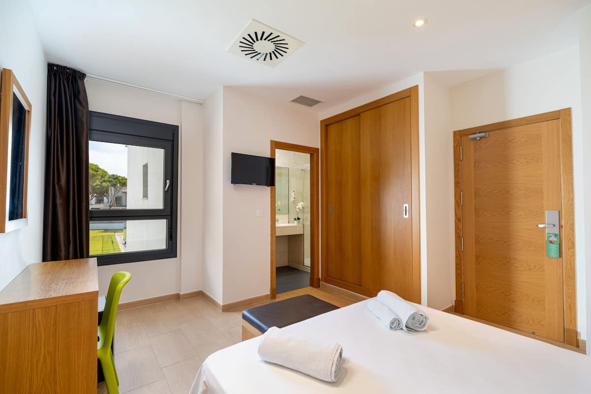 Habitación de hotel con cama, escritorio, silla, televisor, armario y baño visible a través de una puerta abierta.