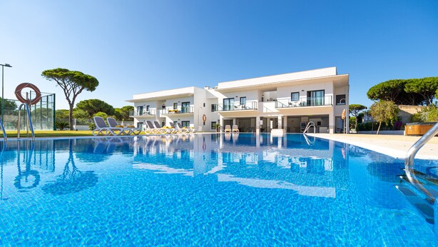 Una piscina con agua cristalina azul, rodeada de edificios modernos con balcones y un árbol.