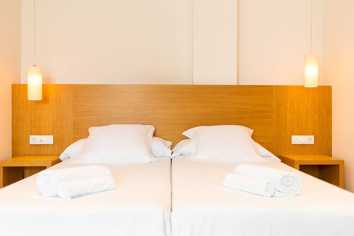 Cama doble con sábanas blancas y dos almohadas, cabecera de madera, mesitas de noche con lámparas y toma de corriente a cada lado.