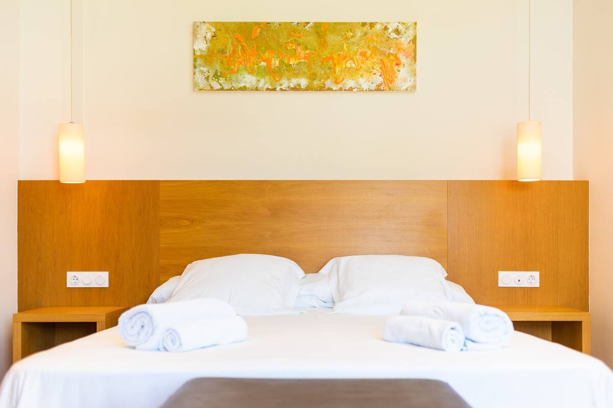 Una cama bien hecha con sábanas blancas y toallas enrolladas, un cabecero de madera y dos mesitas de noche con lámparas.