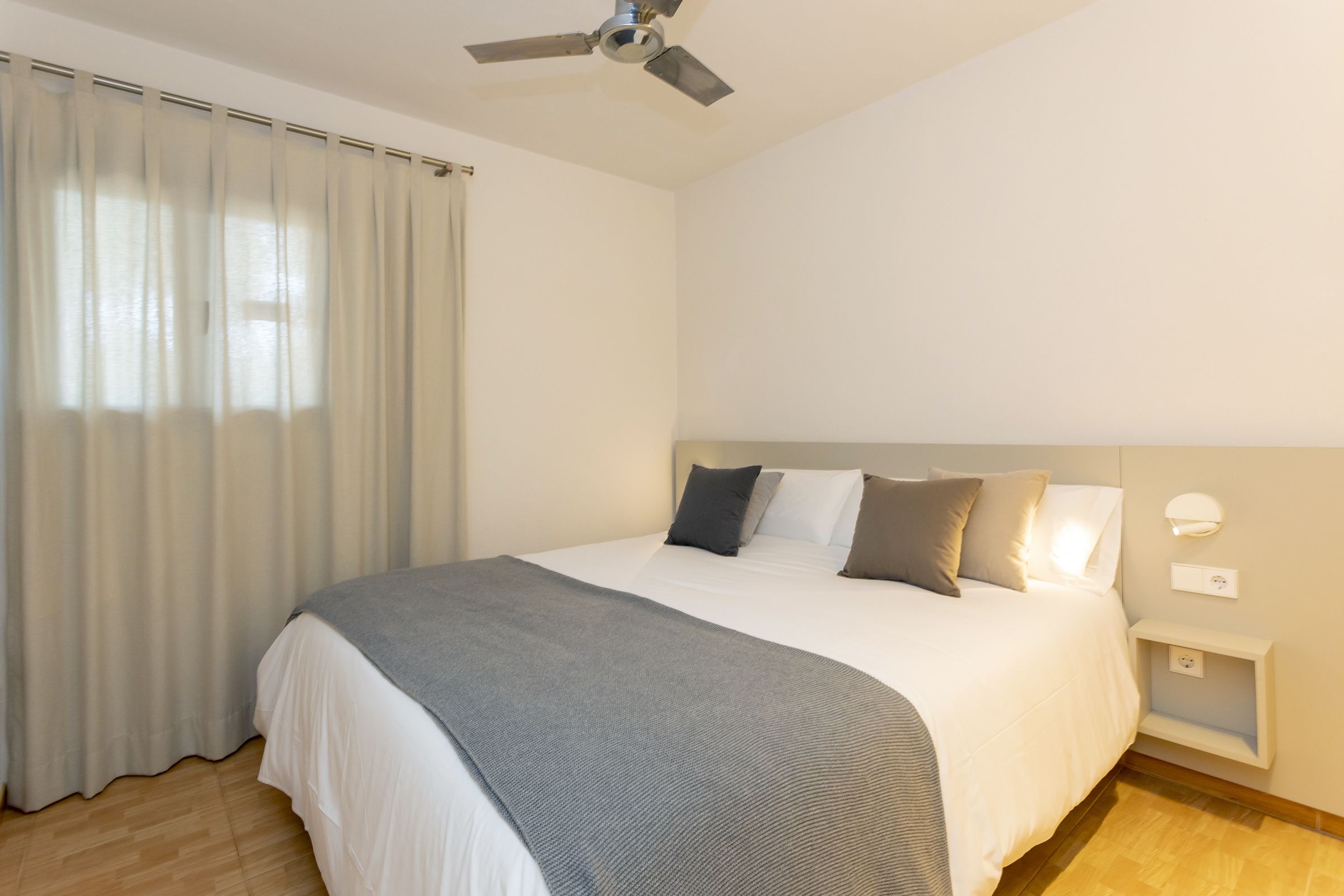 A neatly made bed with a grey blanket, a ceiling fan, and a bedside table with a lamp.