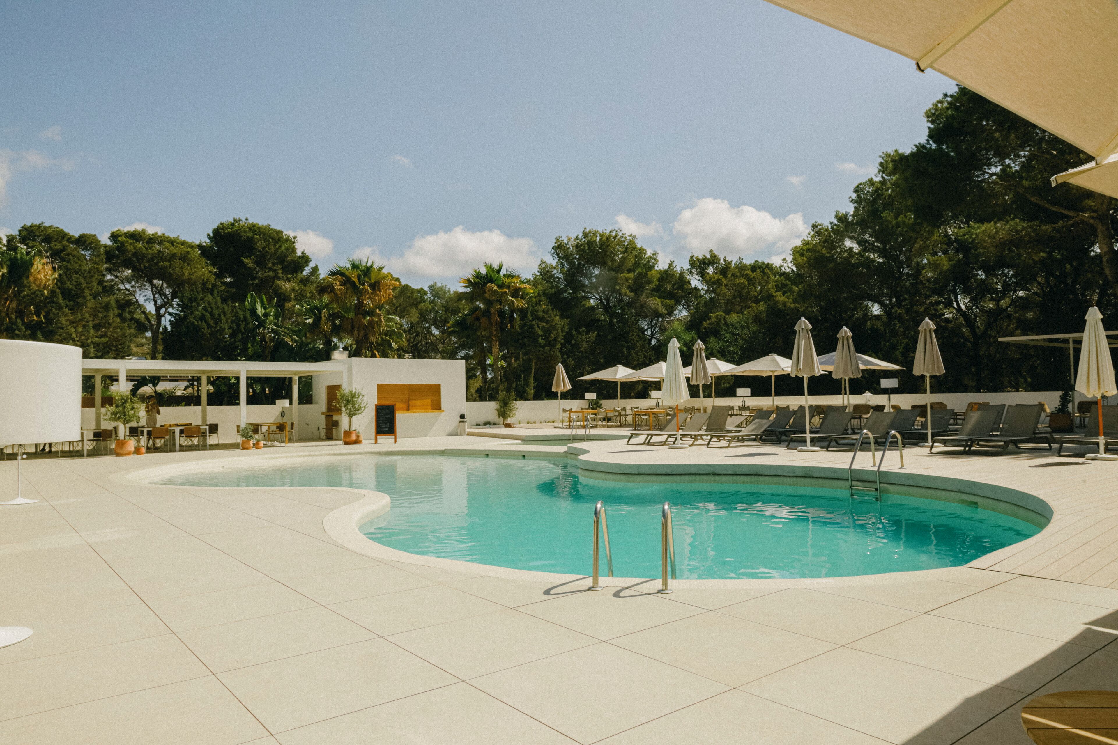 A swimming pool with lounge chairs and umbrellas, surrounded by trees and a building with outdoor seating.