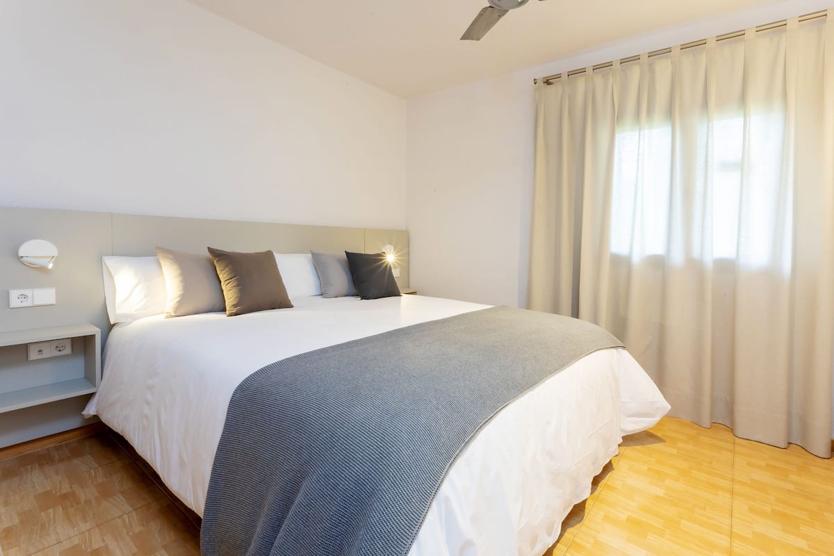 Ein ordentlich gemachtes Bett mit grau-weißer Decke, einem Kopfteil und einem Nachttisch mit Lampe.