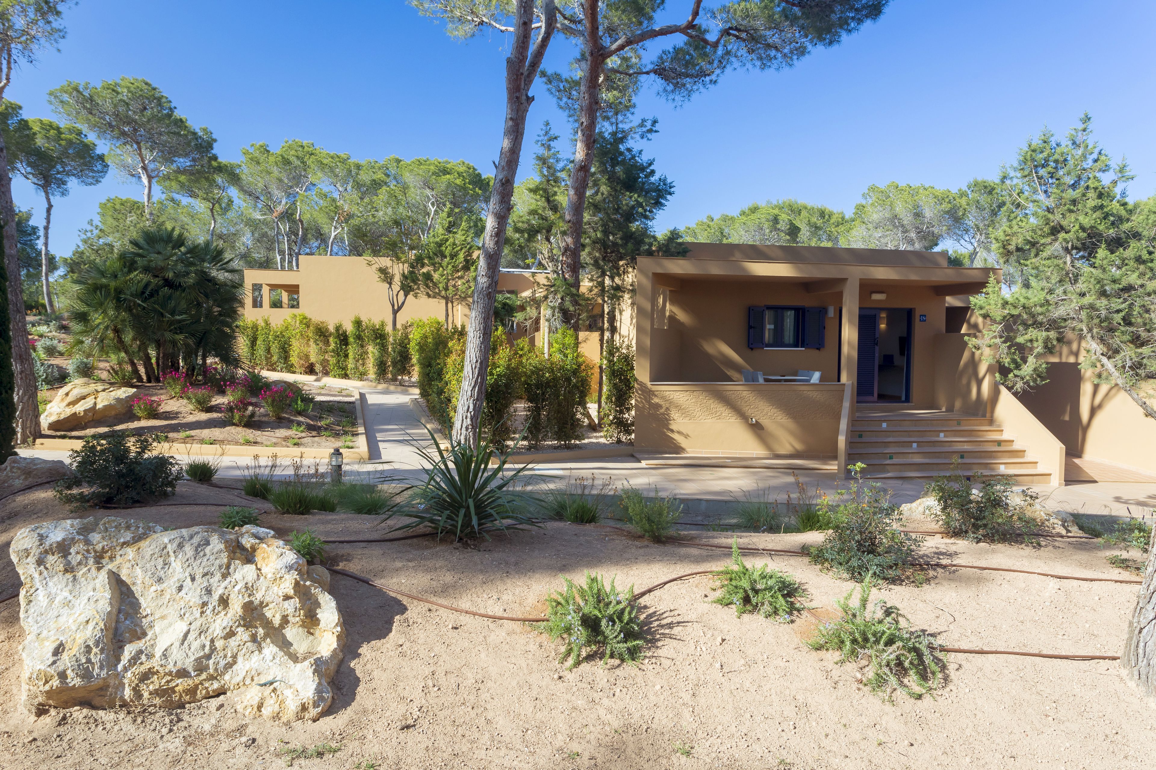 Un edificio moderno con facciata beige, circondato da un giardino curato con diverse piante e un grosso masso in primo piano.