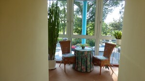 A room with a large window, wicker chairs, a round table, and potted plants. - Hotel Alpha (Nuremberg)