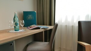 A hotel room with a wooden desk, a grey chair, a bottle of Sodena, two wine glasses, and a magazine. - Hotel Alpha (Nuremberg)