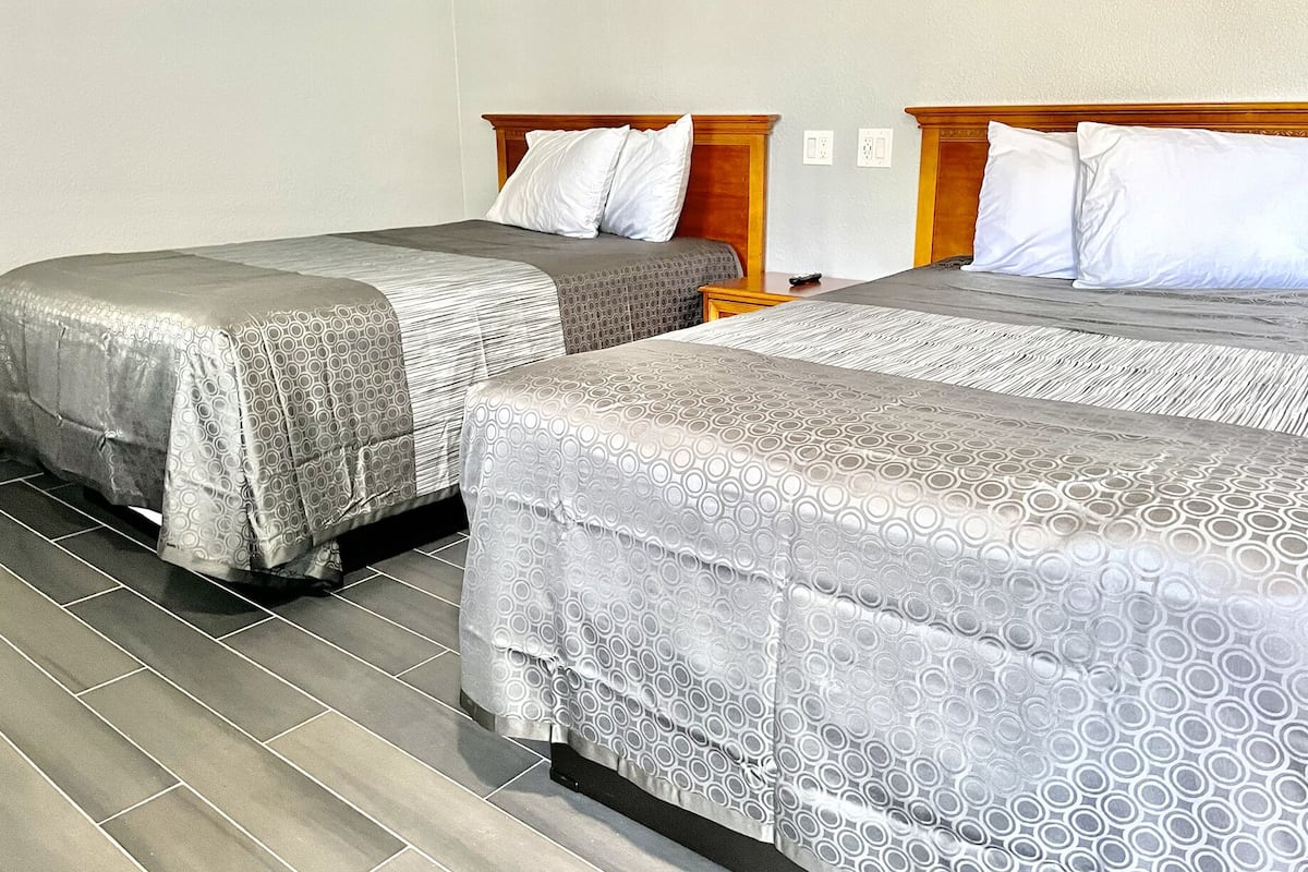 Two single beds with grey patterned bedspreads, wooden headboards, and white pillows in a room with grey tiled flooring.