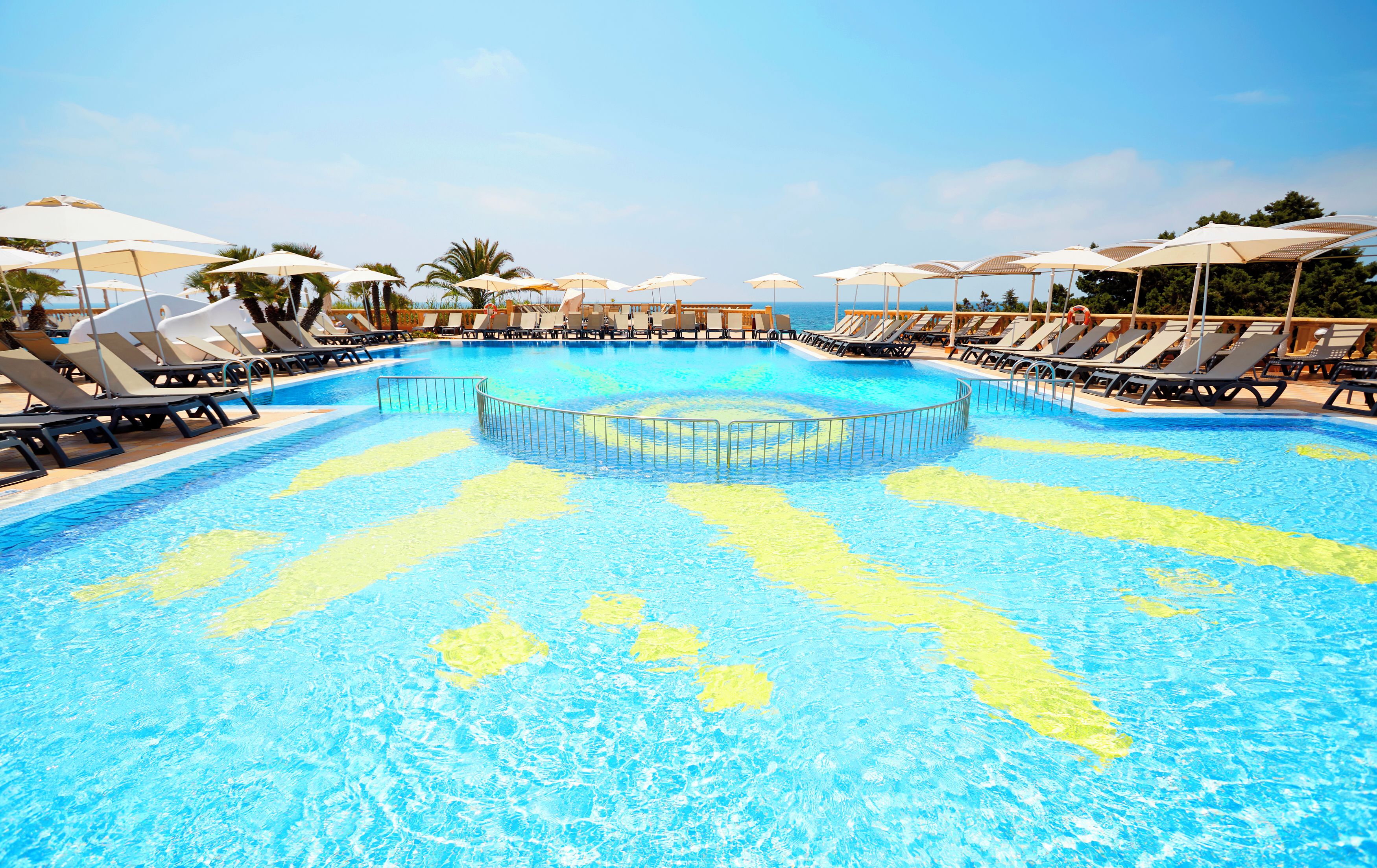 A large pool with multiple sun loungers and umbrellas, surrounded by palm trees and a clear sky.