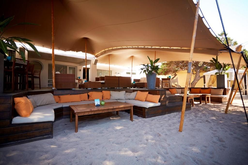 A covered outdoor seating area with sofas and a wooden table, surrounded by sand.