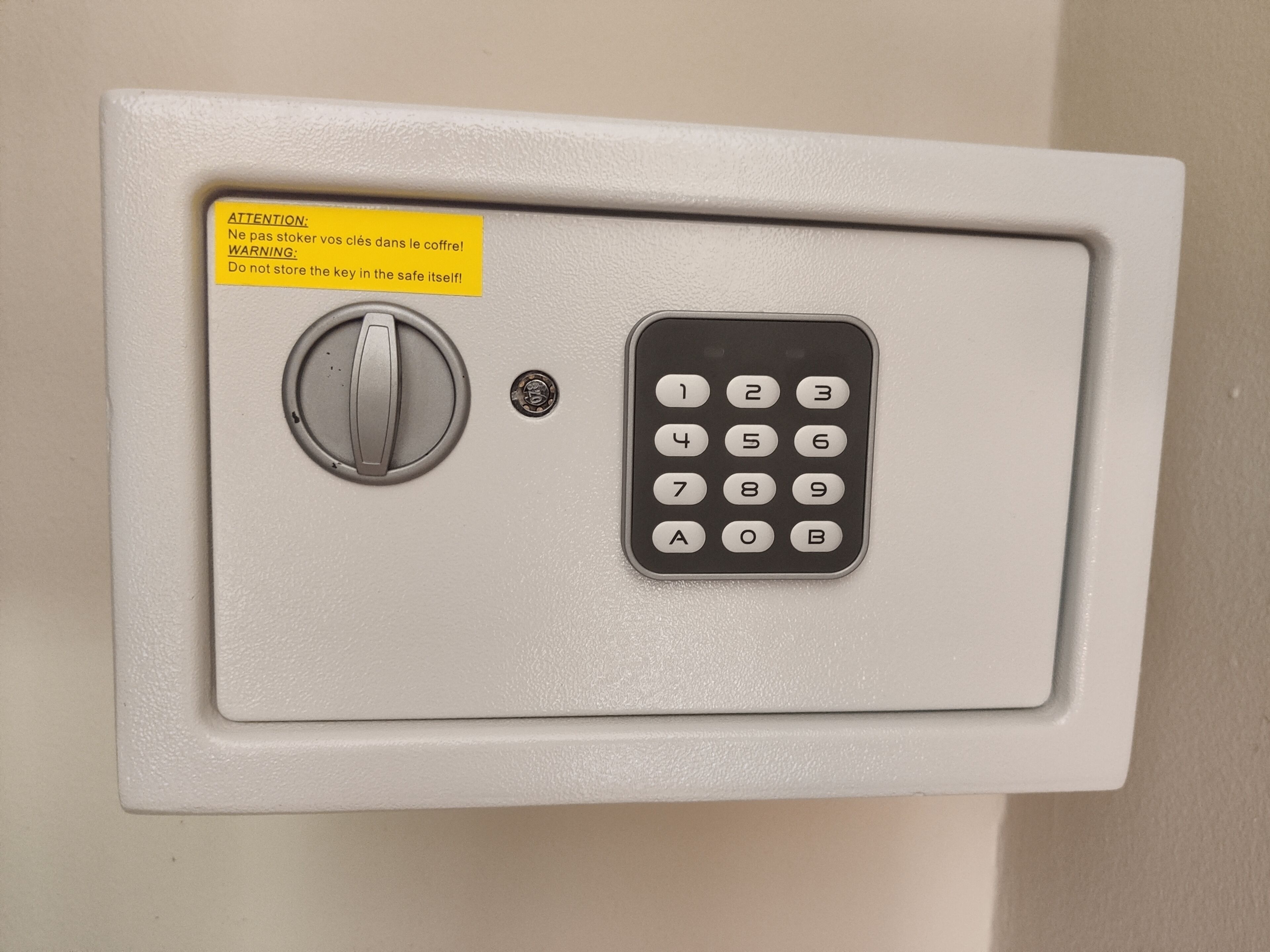 A safe deposit box with a numeric keypad and a keyhole, featuring a warning label in French advising not to store the key inside the safe.