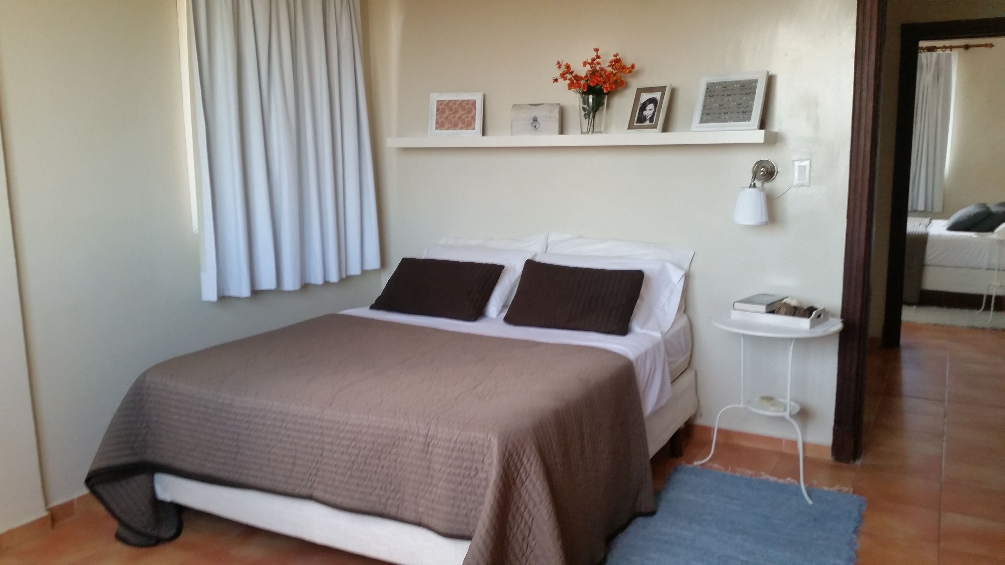 A bedroom with a bed, a bedside table, a nightstand, a wall-mounted shelf with pictures and a vase of flowers, and a window with curtains.