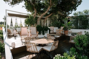 An outdoor patio with wicker furniture, a round table, and a large tree providing shade. - Hotel Rosamar (Formentera)