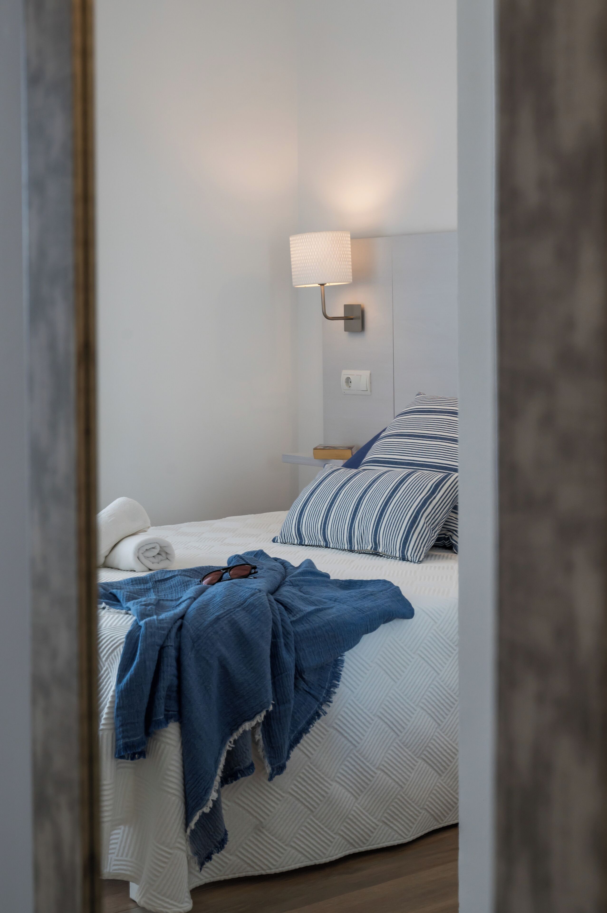 Una cama bien hecha con un cojín azul y blanco a rayas, una manta azul y un par de gafas de sol sobre la cama.