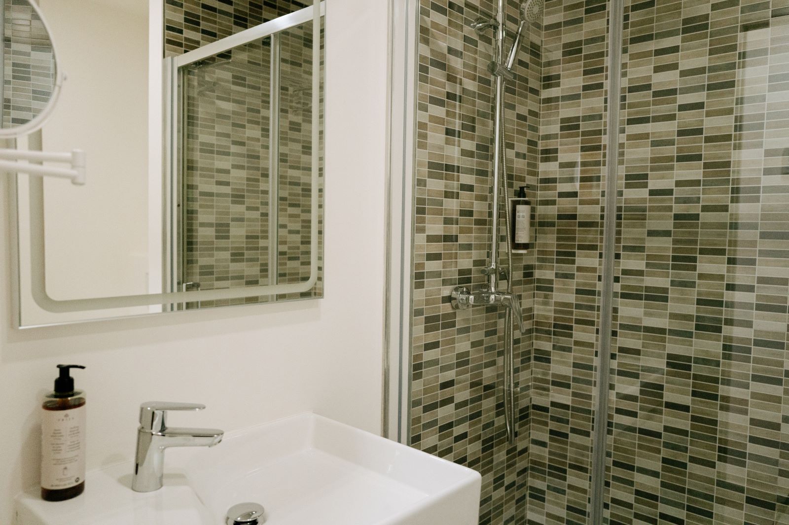 Un baño con lavamanos blanco, un espejo, ducha con puerta de cristal y pared con azulejos.