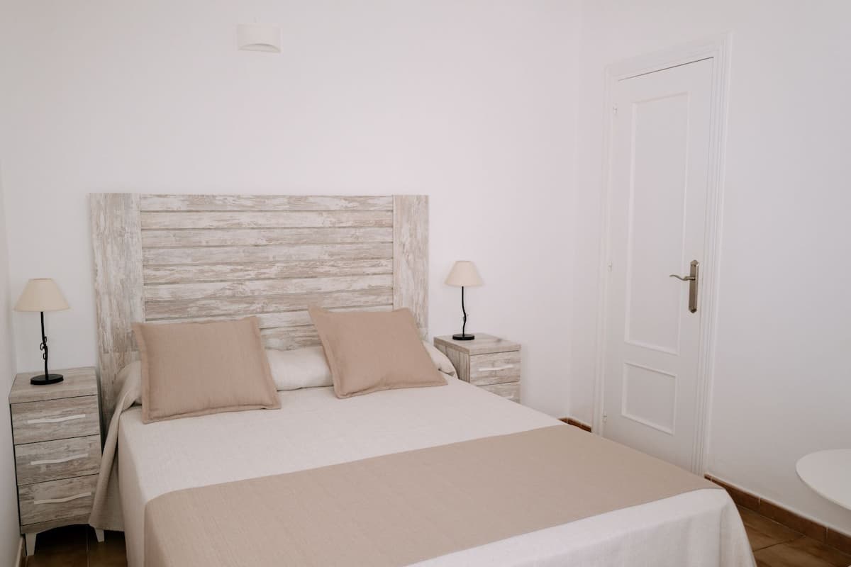 A bedroom with a bed, two bedside tables, a nightstand, a door, and a wall-mounted light fixture.