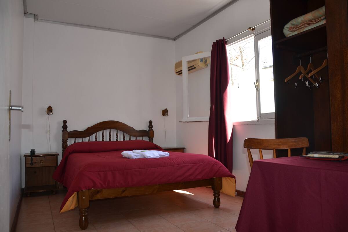 A bedroom with a wooden bed, a nightstand, a table with a red tablecloth, and a window with curtains.
