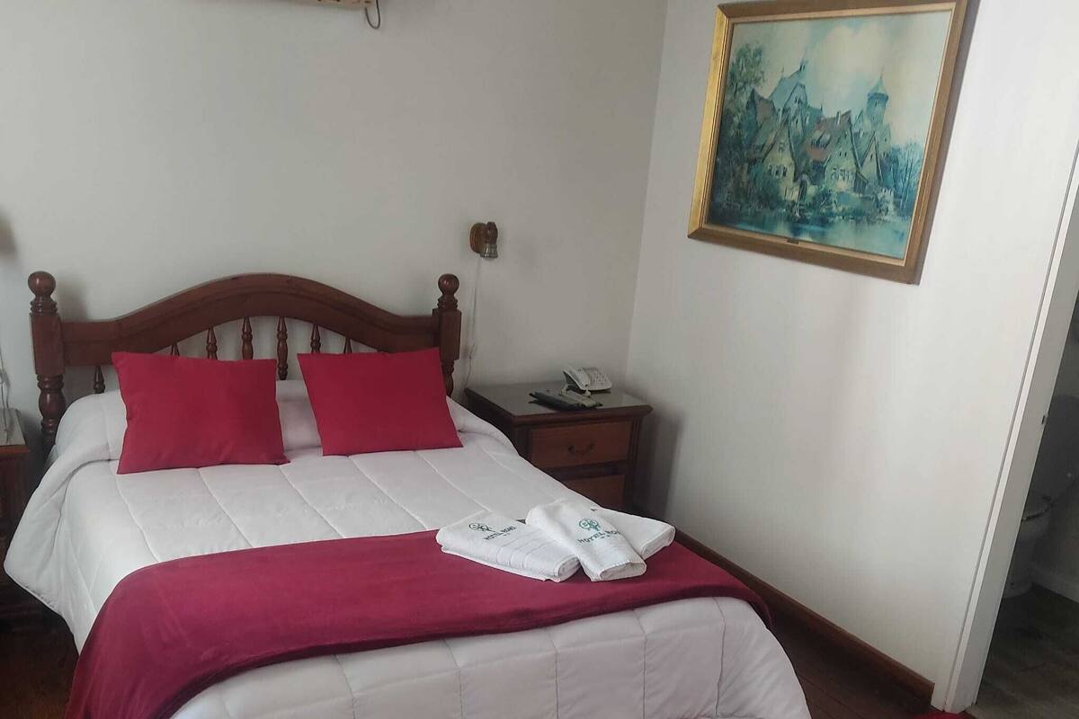 A neatly made bed with red pillows and a white duvet, a wooden nightstand with a phone, and a painting on the wall.