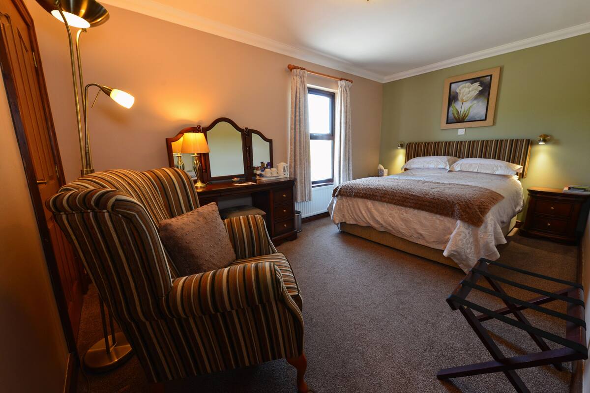 A hotel room with a bed, a chair, a desk with a lamp, a window with curtains, and a painting on the wall.