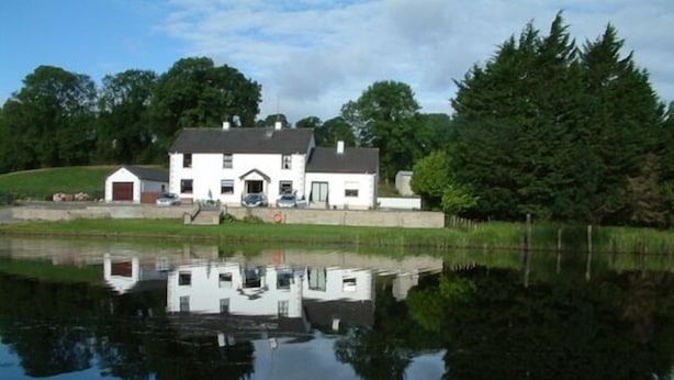A white house with a garage and a small shed, surrounded by trees and a pond. - Corrigans Shore House (Enniskillen)