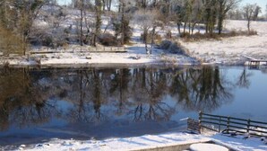 A snow-covered outdoor area with a wooden table and benches, a pond, and trees. - Corrigans Shore House (Enniskillen)