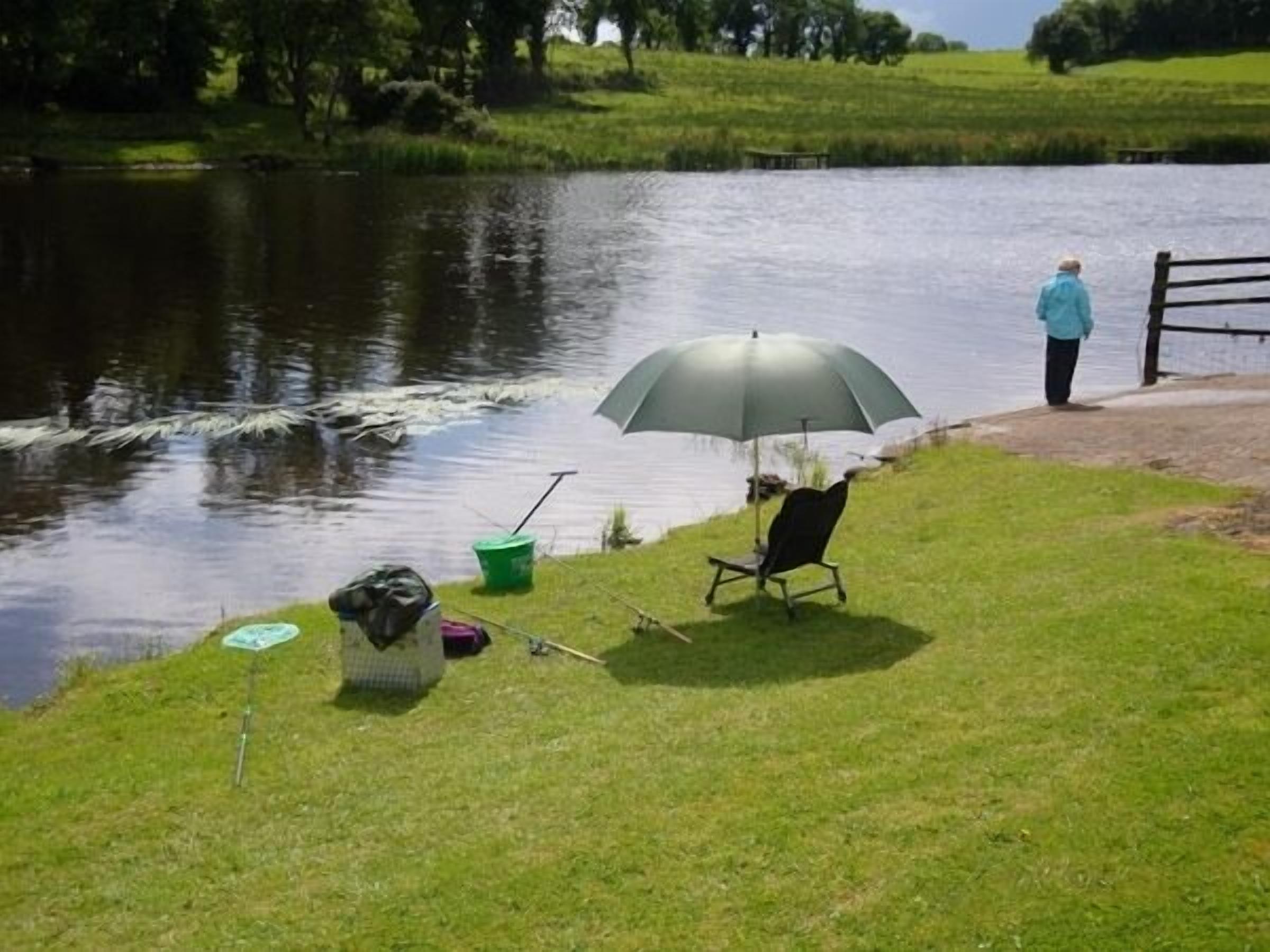 A lakeside setting with a chair under an umbrella, a green bucket, and a bag on the grass.