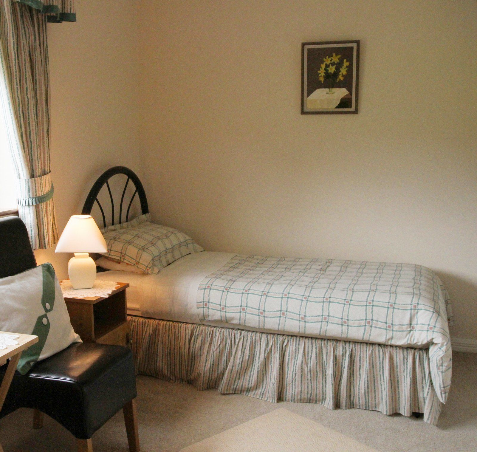 Un dormitorio con una cama individual, una silla, una mesita de noche, una lámpara, un cuadro en la pared y una ventana con cortinas.