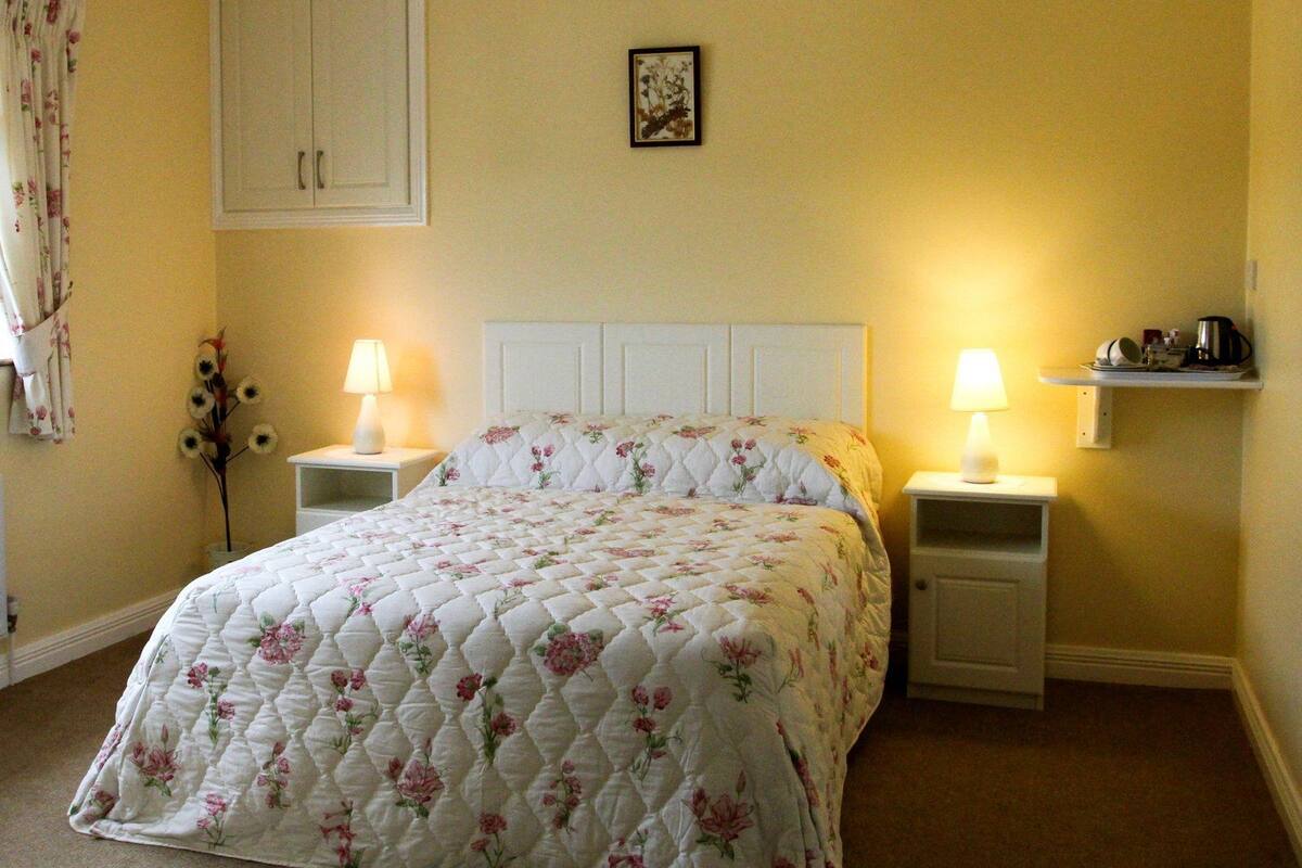 A neatly made bed with a floral quilt, two bedside lamps, a small shelf with a coffee maker, and a framed picture on the wall.