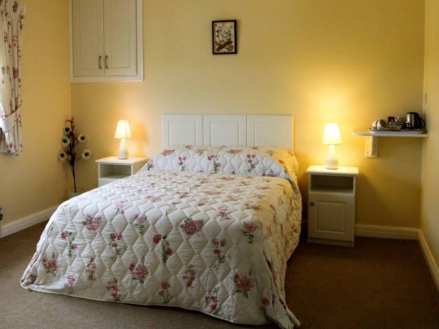 Een keurig opgemaakt bed met een bloemenquilt, twee bedlampjes, een klein plankje met een koffiezetapparaat, en een lijst met een foto aan de muur.