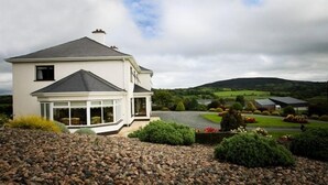 A two-story house with a large bay window and a covered porch, surrounded by a landscaped garden and a gravel driveway. - The Arches (Arvagh)