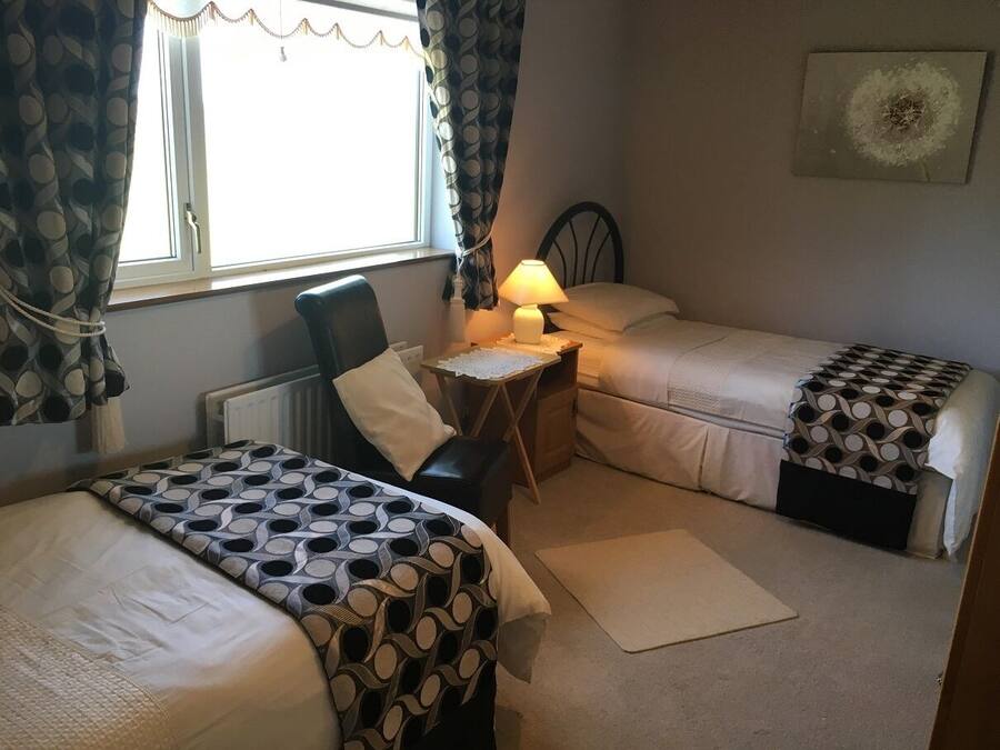 A bedroom with two beds, a chair, a small table with a lamp, and a window with curtains.