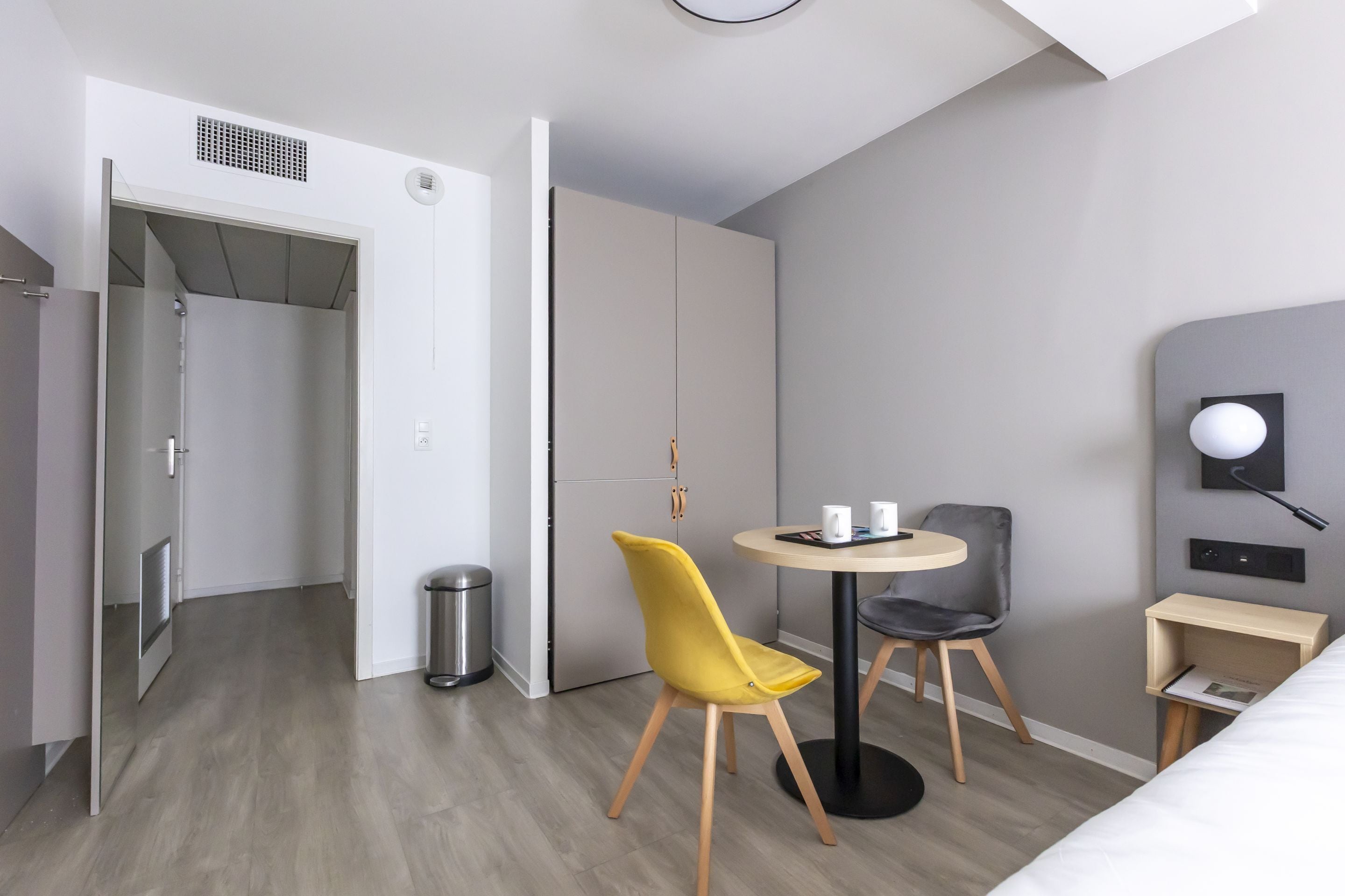 Een moderne hotelkamer met een bed, een klein rond tafeltje met twee stoelen, een bureau met stoel, een kledingkast en een prullenbak.