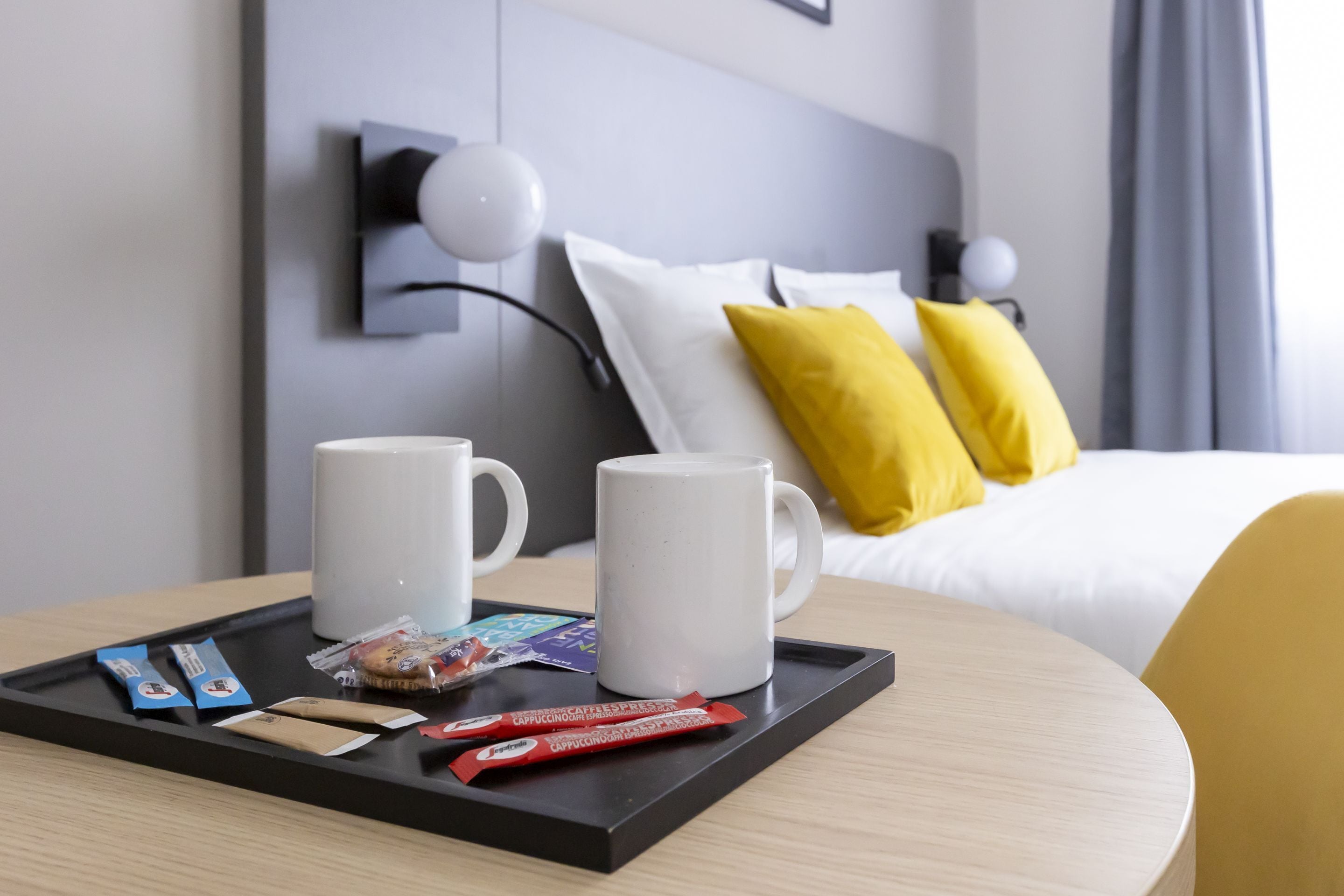Habitación de hotel con una cama, dos almohadas, una bandeja con tazas de café y aperitivos, y una mesita de noche.