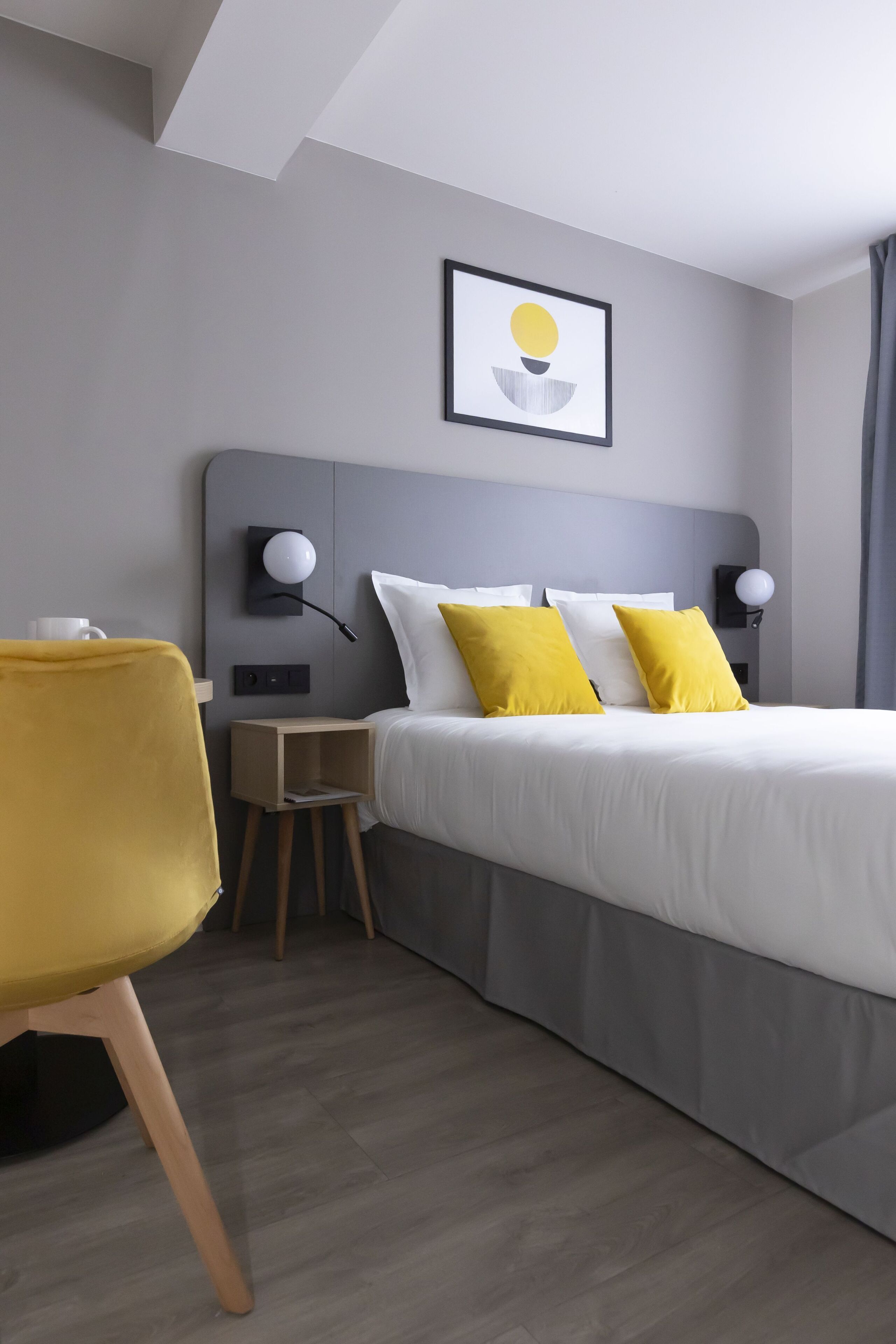 A modern hotel room with a bed, two bedside lamps, a small wooden nightstand, a yellow chair, and a framed picture on the wall.