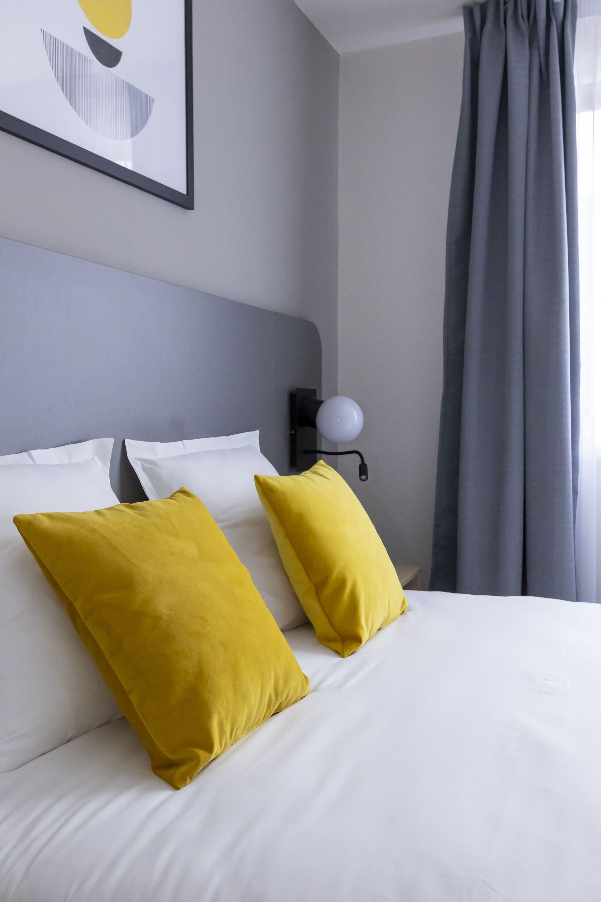 Une chambre d’hôtel avec un lit, deux oreillers, une lampe fixée au mur et une fenêtre avec des rideaux.