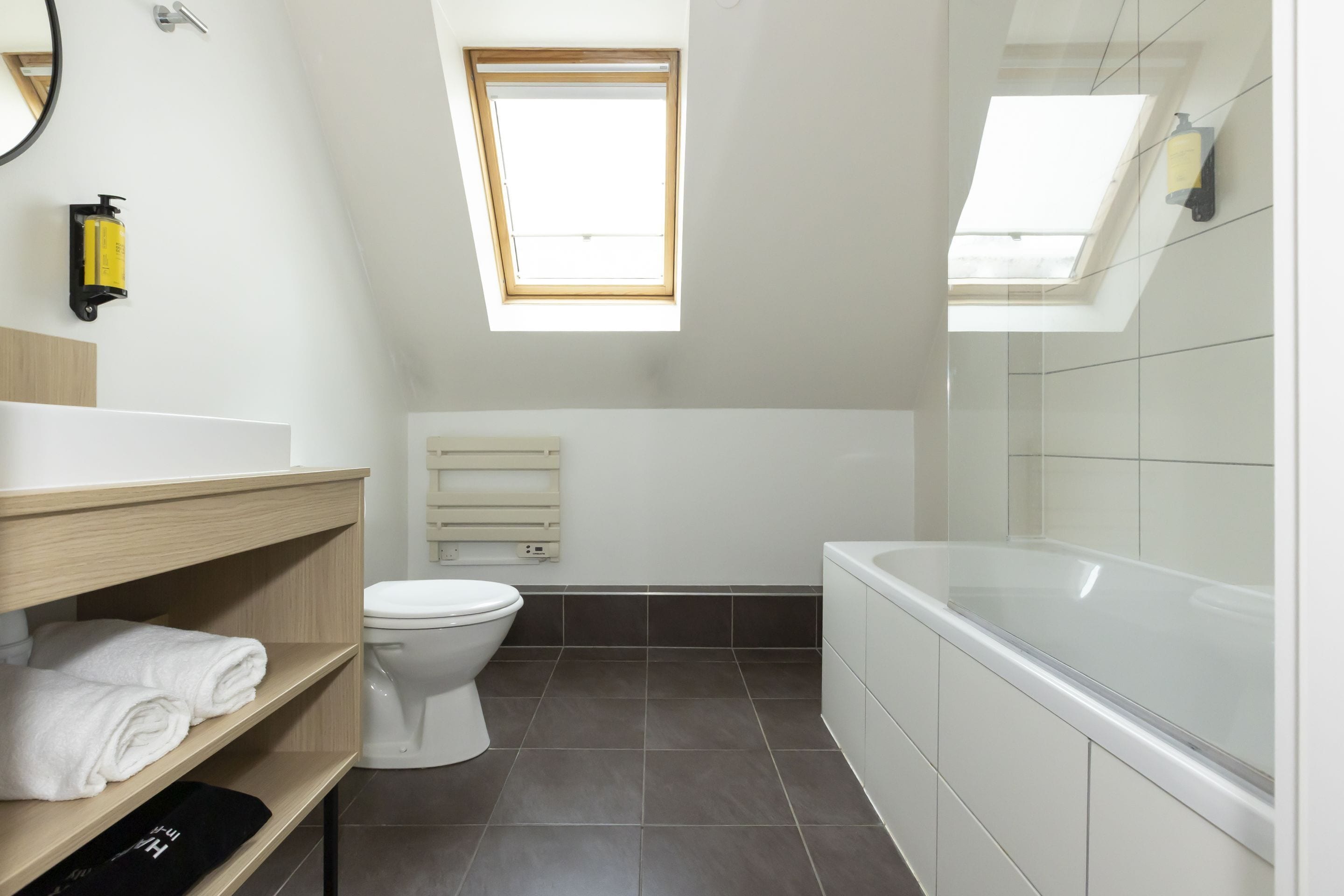 A modern bathroom with a skylight, a bathtub, a toilet, and a shelf with towels.