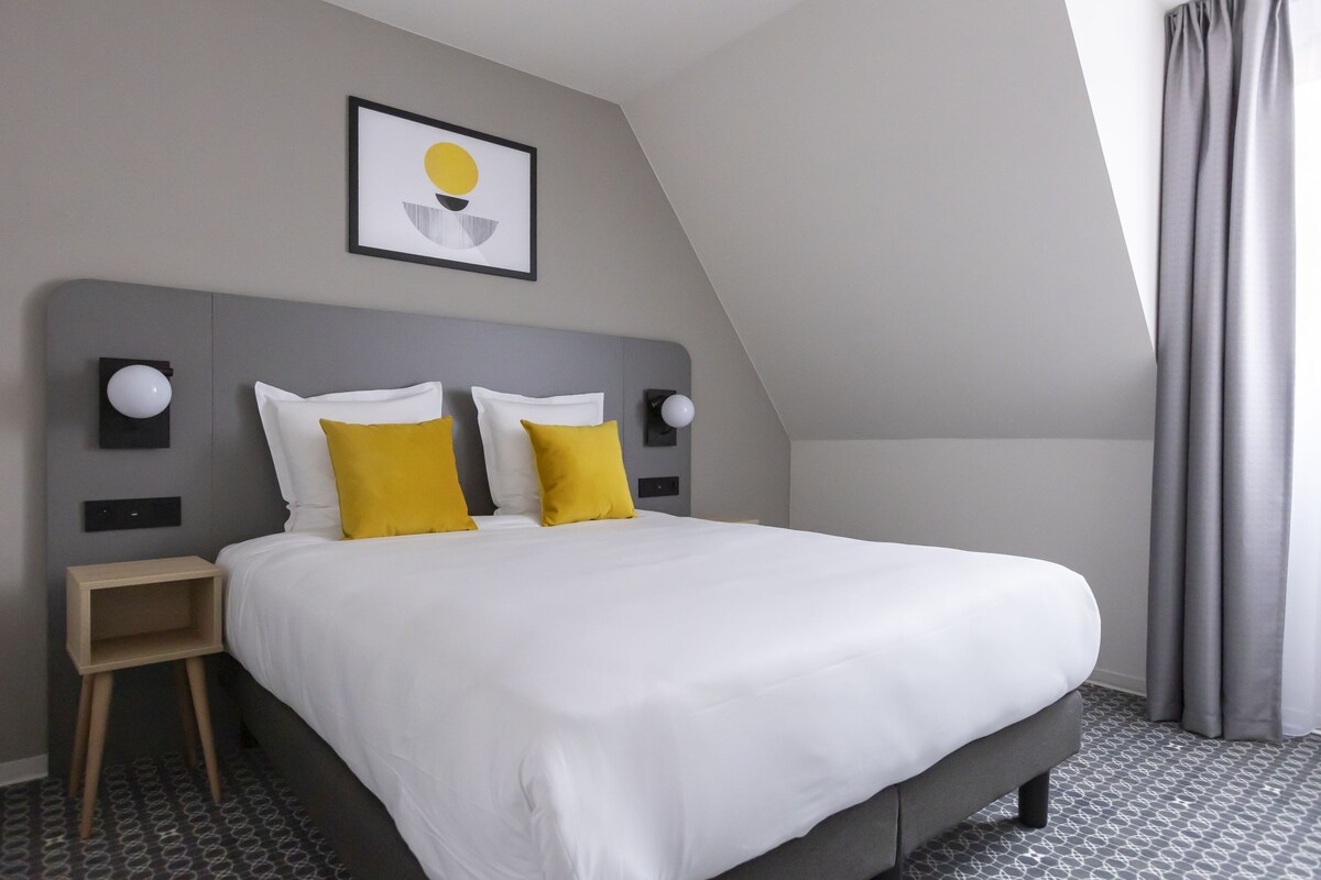 Quarto de hotel moderno com uma cama grande, duas mesinhas de cabeceira, uma luminária na parede e um quadro emoldurado sobre a cama.