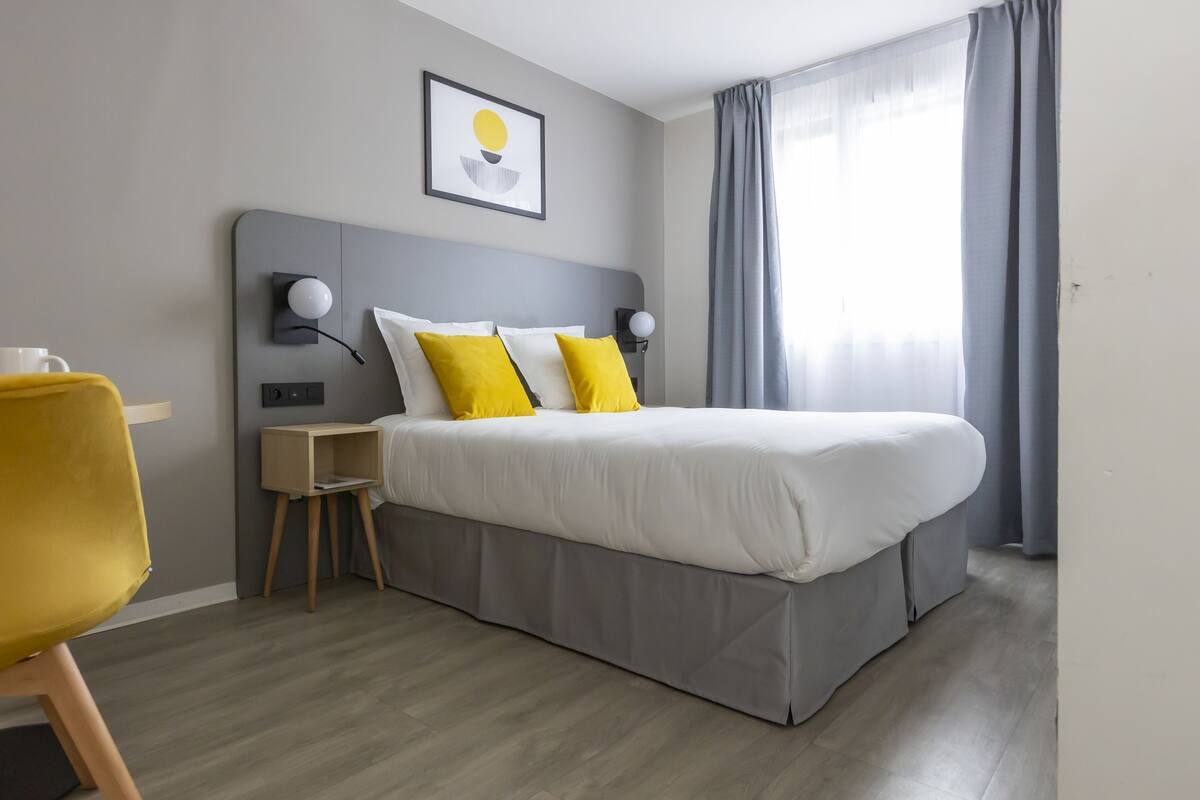 A modern hotel room with a large bed, two bedside lamps, a small wooden nightstand, a yellow chair, and a framed artwork on the wall.