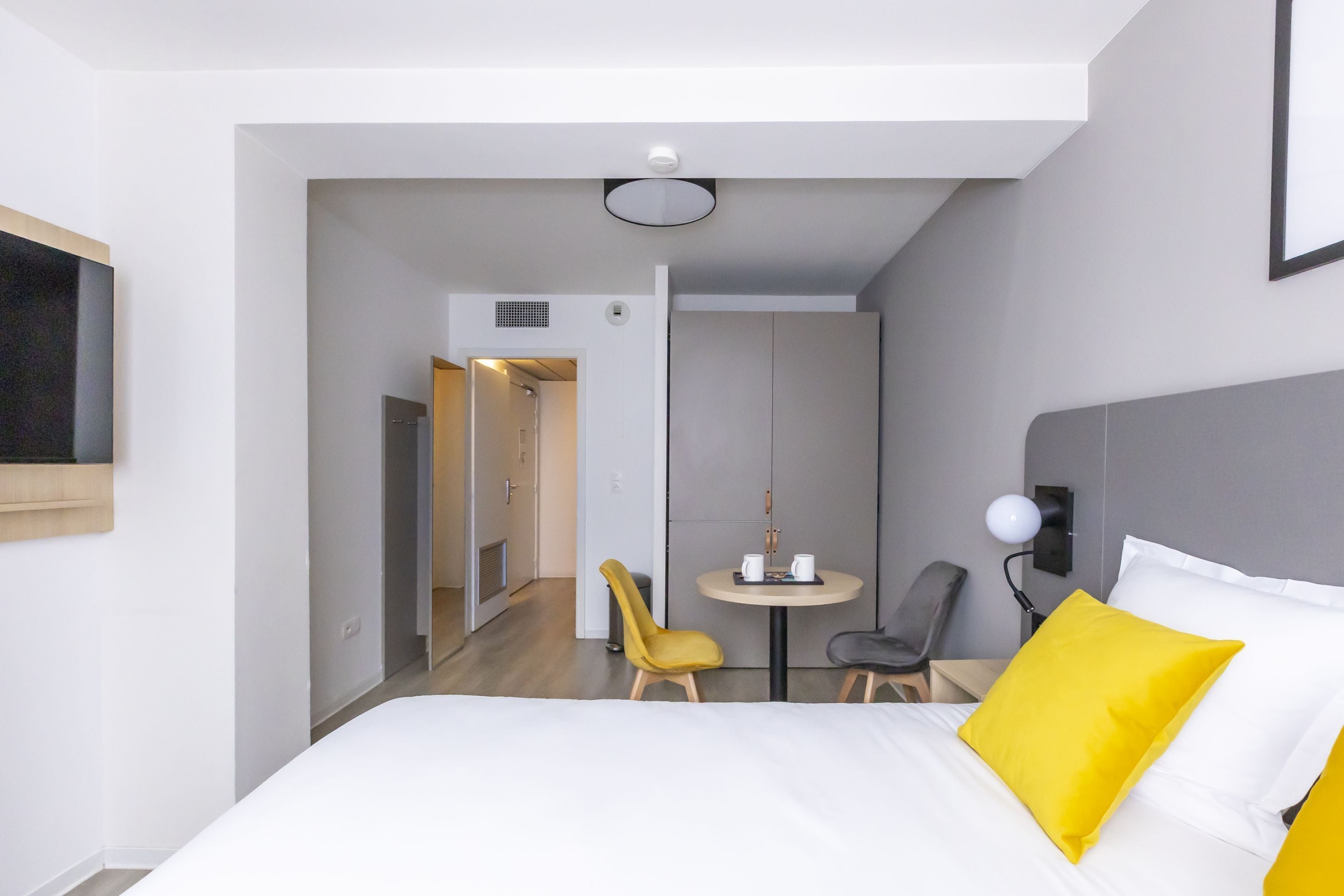A modern hotel room with a bed, a small table with chairs, a TV, and a door leading to another room.