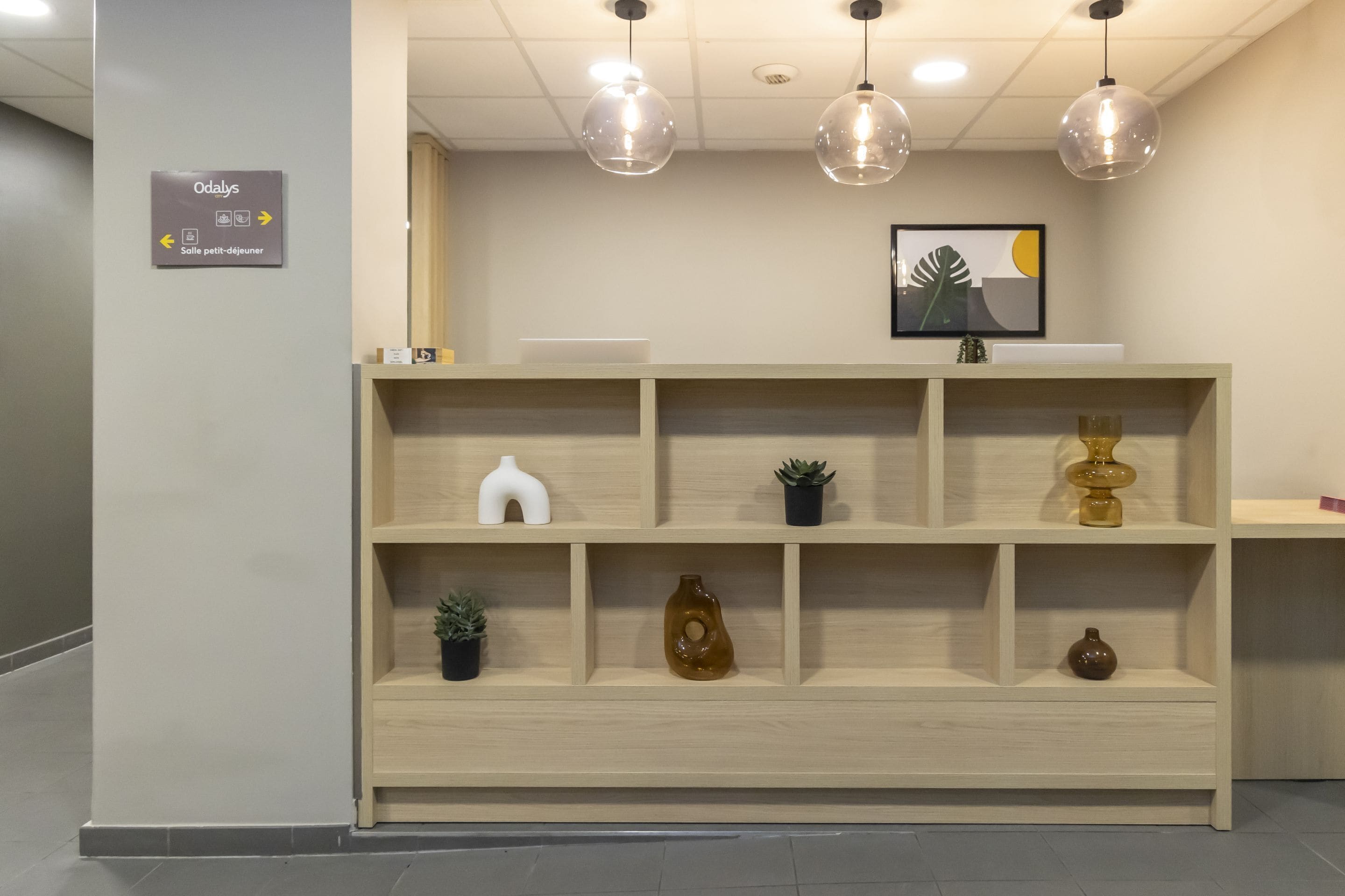 Un área de recepción con un cartel de Odalys, un espacio tipo apartamentos, y un banco de madera con elementos decorativos.