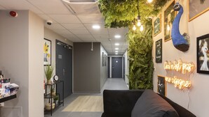 Un espace d’accueil moderne avec un canapé noir, un comptoir d’accueil où sont disposées des brochures, un panneau lumineux au néon et des décorations murales, notamment un paon et un oiseau.