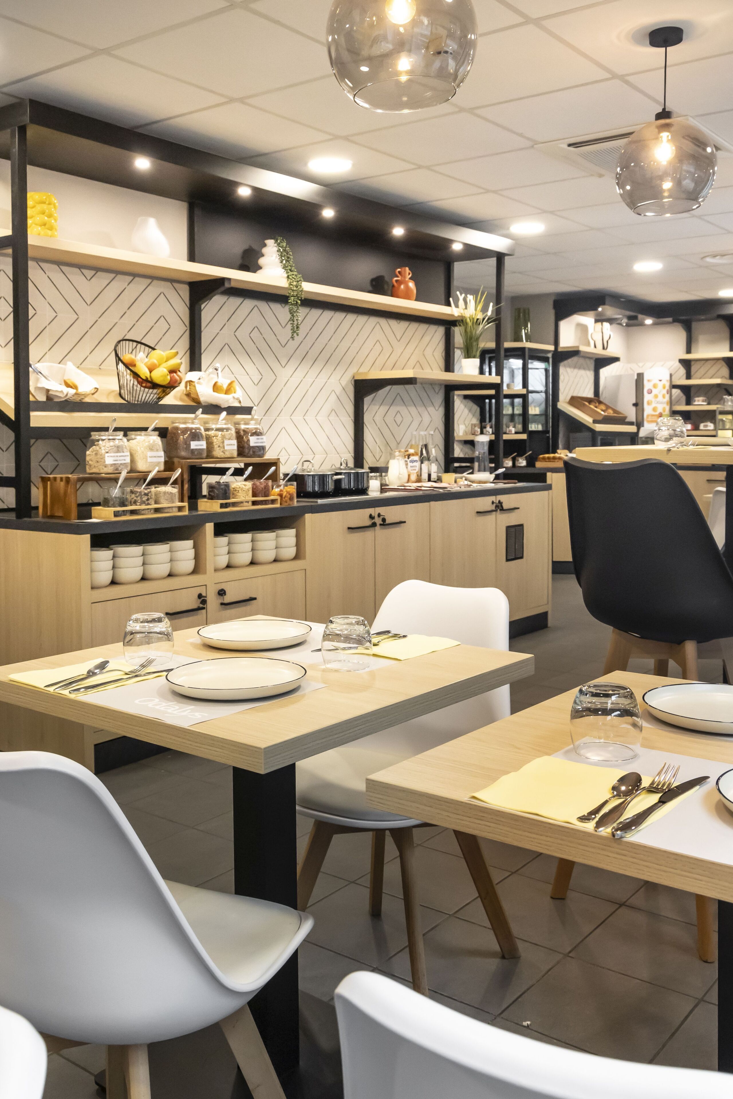 A modern dining area with wooden tables and chairs, a kitchen in the background, and pendant lighting.