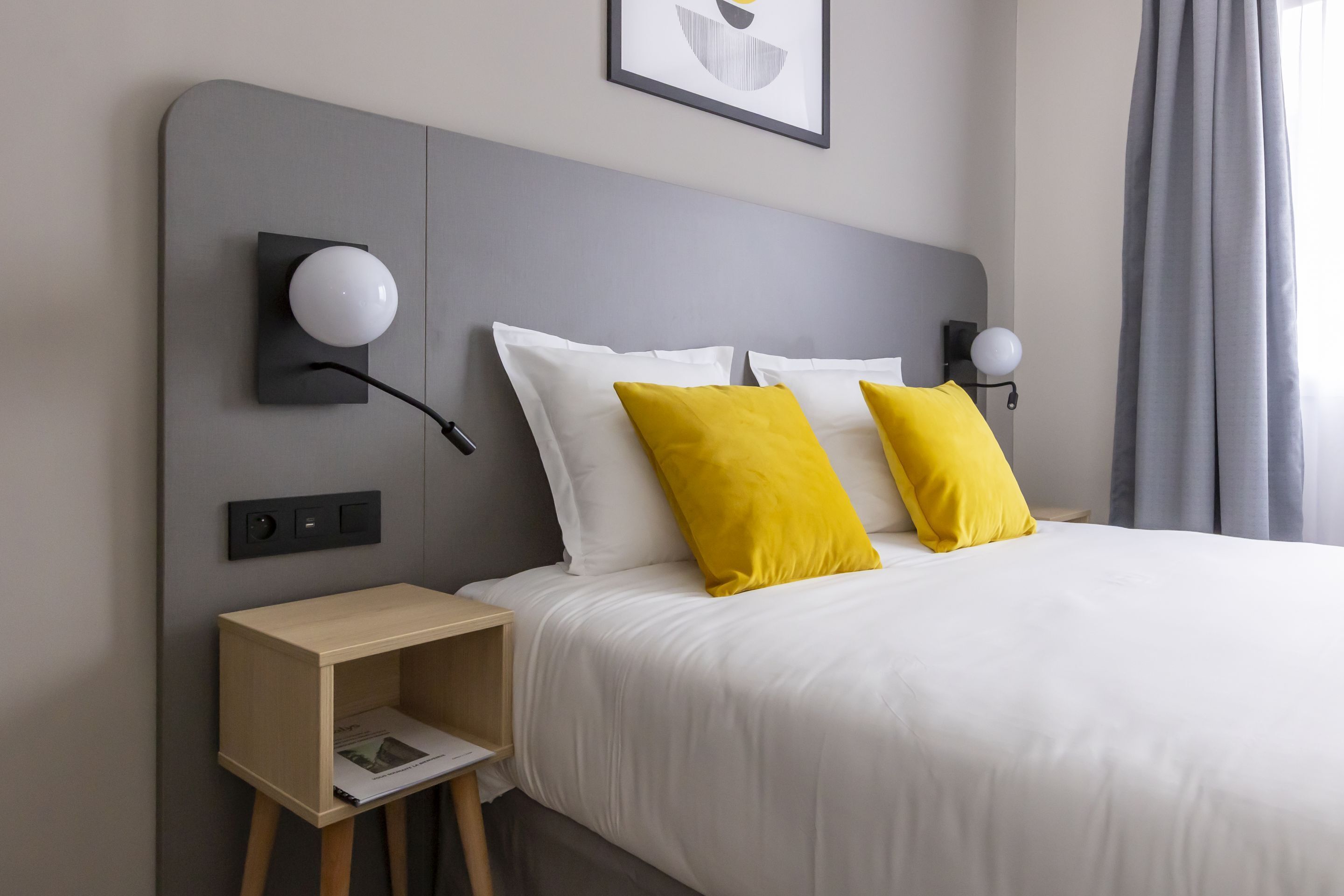 Une chambre d’hôtel moderne avec une tête de lit grise, du linge de lit blanc et deux oreillers jaunes.