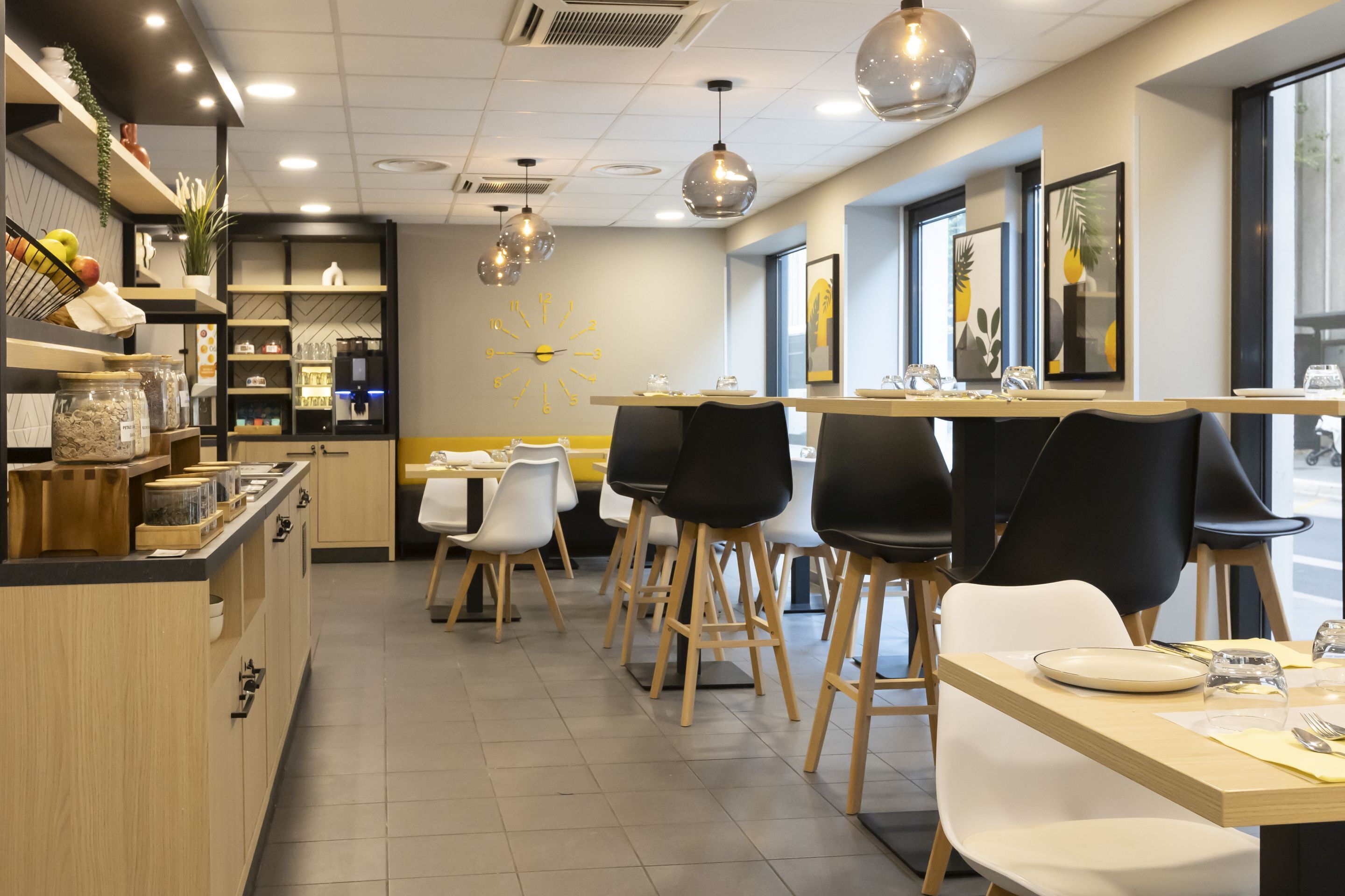 Un intérieur de restaurant moderne avec des tables et des chaises en bois, un espace cuisine doté d’étagères et de bocaux, et une grande horloge murale.