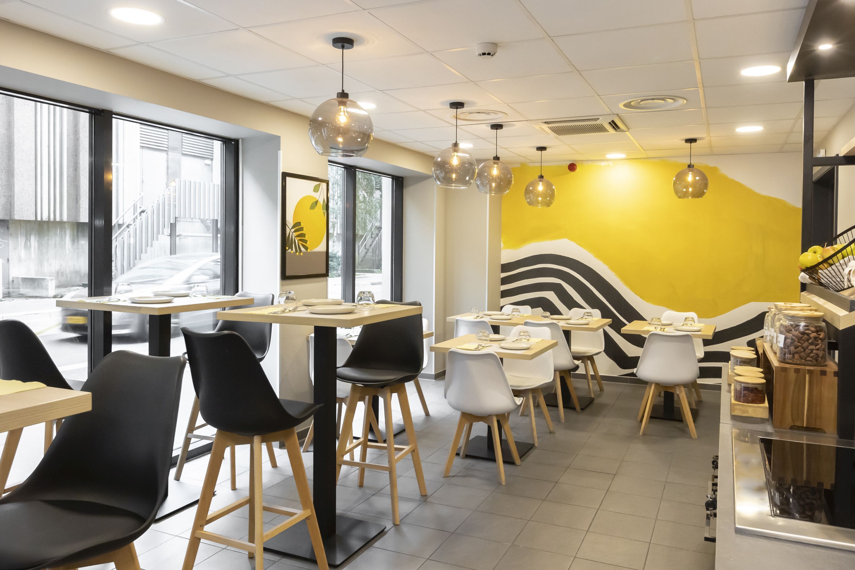 Interior de una cafetería moderna con mesas y sillas, un mural amarillo y lámparas colgantes.