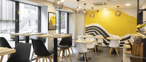 Interior de una cafetería moderna con mesas y sillas, un mural amarillo y lámparas colgantes.