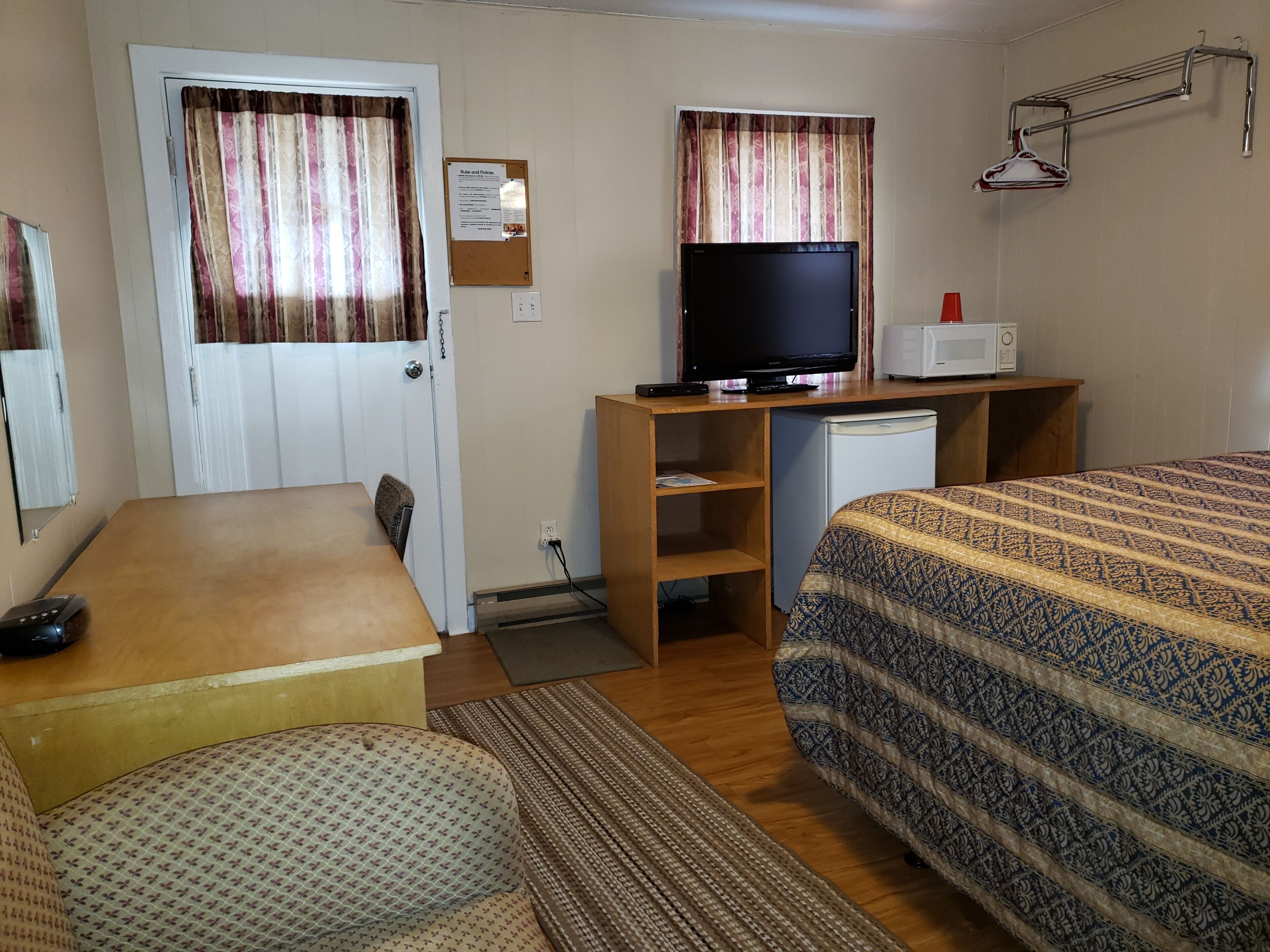 Une chambre d’hôtel de taille réduite, équipée d’un lit, d’un bureau avec un ordinateur, d’une télévision et d’un four à micro-ondes.