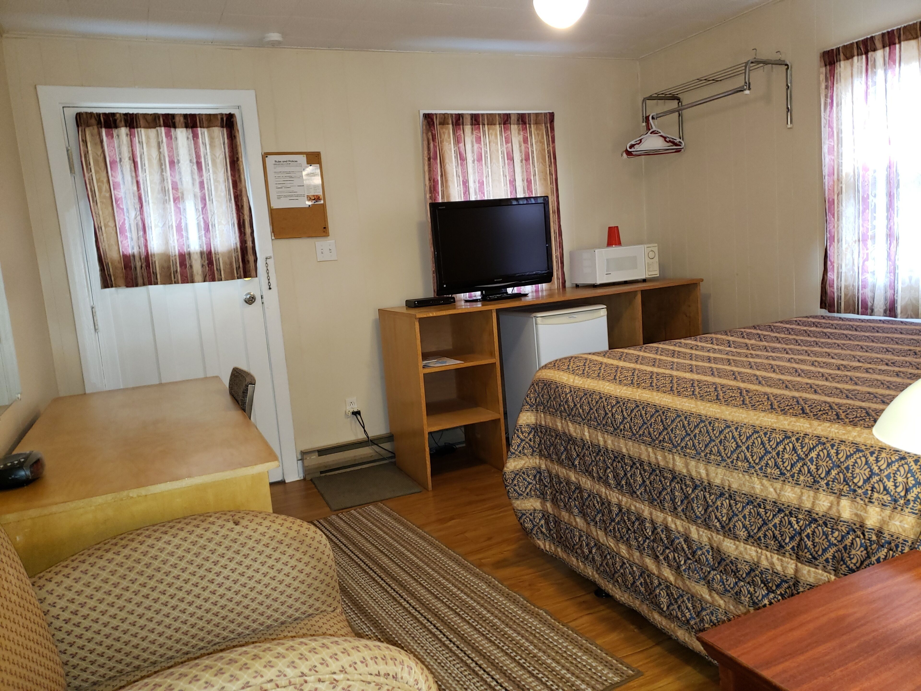 A hotel room with a bed, a desk with a television, a small refrigerator, and a seating area with a sofa and armchair.