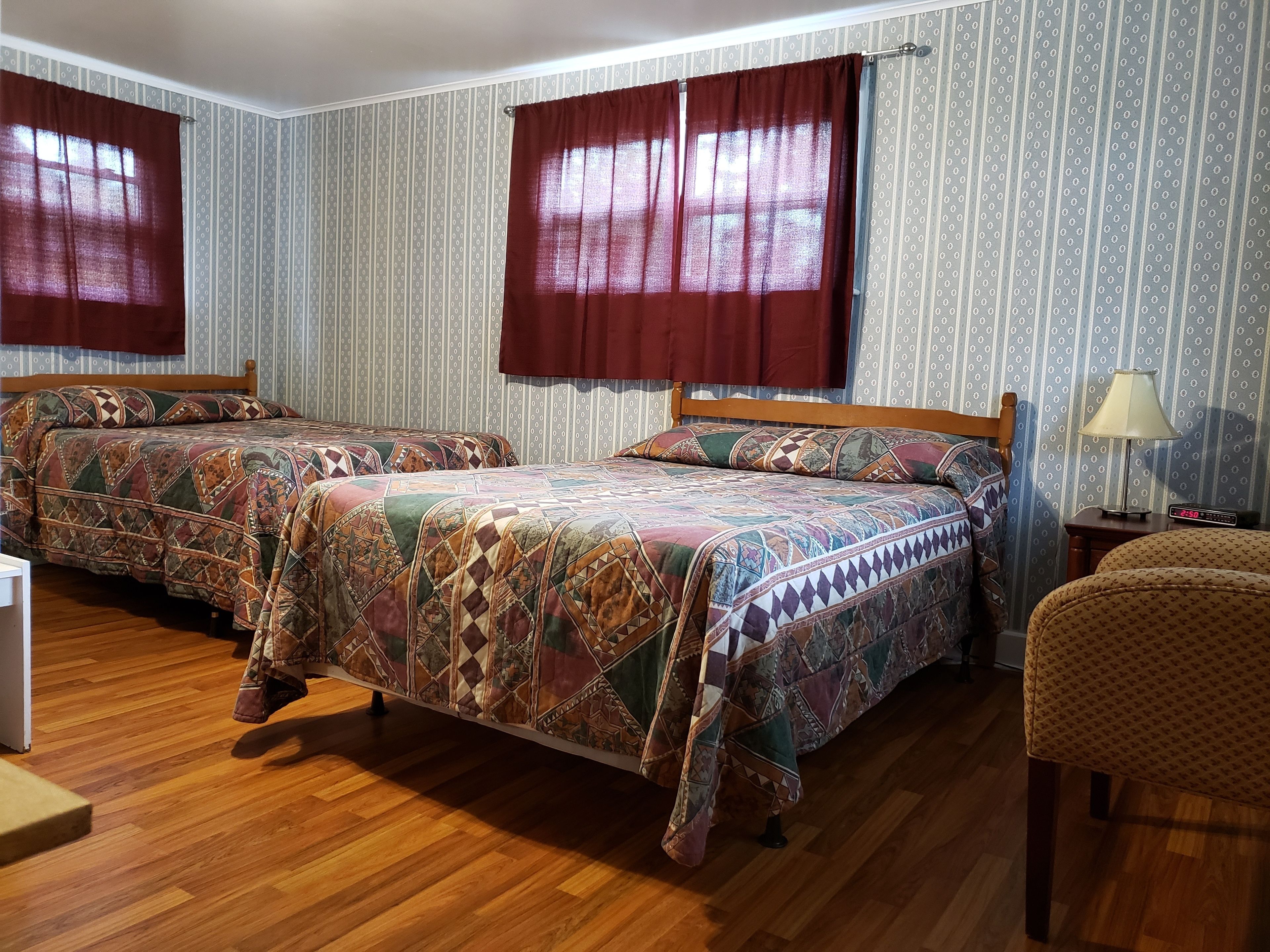 Una habitación con dos camas, piso de madera, papel tapiz estampado y una mesita de noche con lámpara.